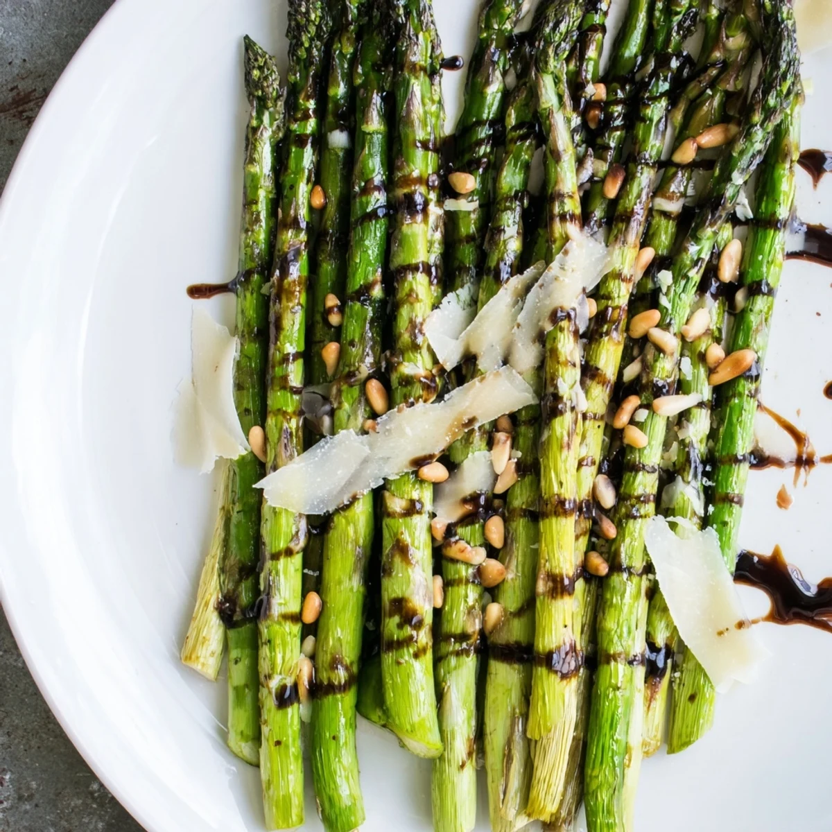 Roasted Asparagus with Balsamic Glaze on a white platter garnished with pine nuts and shaved Parmesan cheese.