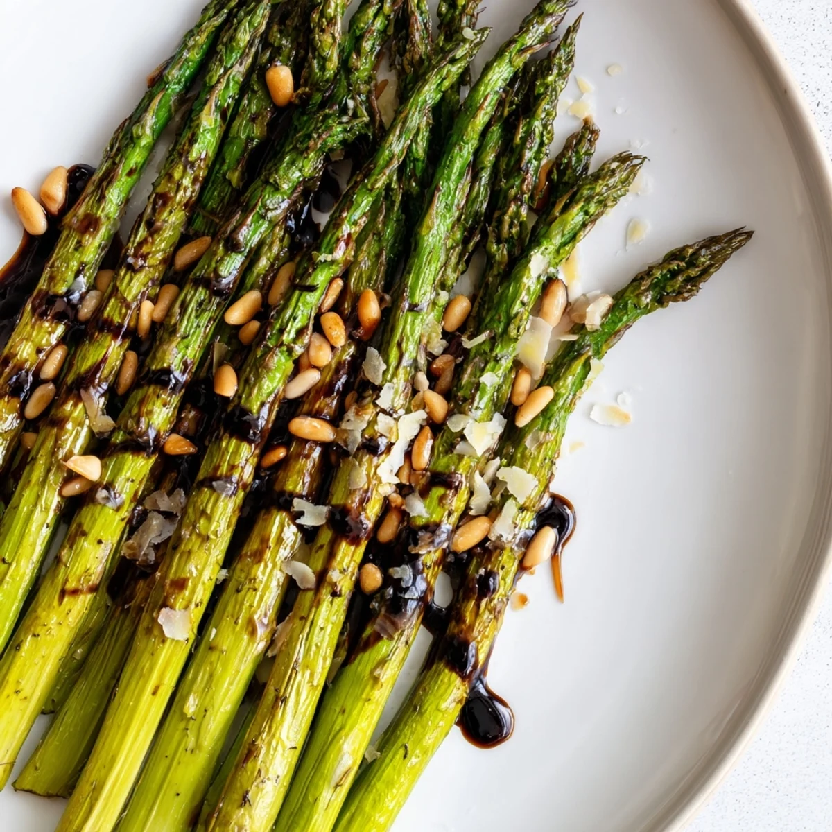 Tender roasted asparagus with balsamic reduction on a white platter, garnished with pine nuts.