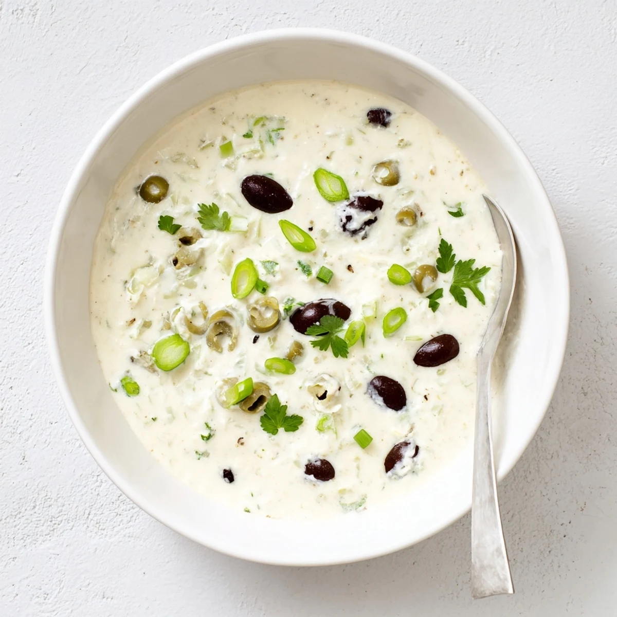 Cream cheese olive dip in white serving bowl surrounded by crackers and fresh vegetable sticks